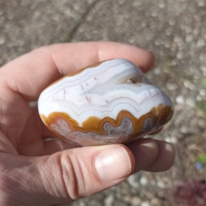 May include: A polished, oval-shaped stone with a natural pattern of white, tan, and brown bands. The stone is held in a hand, showcasing its smooth texture and unique geological formations. The stone's surface has a glossy finish.