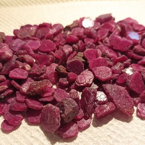 May include: A collection of raw ruby gemstones. The stones are a deep red colour, with variations in size and shape. Some stones have darker, brownish patches. The gemstones are scattered on a light-coloured surface.