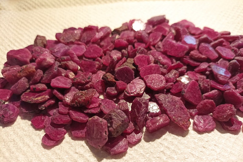 May include: A collection of raw ruby gemstones. The stones are a deep red colour, with variations in size and shape. Some stones have darker, brownish patches. The gemstones are scattered on a light-coloured surface.