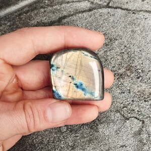 Könnte beinhalten: Ein glatter, polierter Labradorit-Edelstein in hellbraun mit blauen und schwarzen irisierenden Blitzen. Der Stein hat die Form eines abgerundeten Rechtecks.