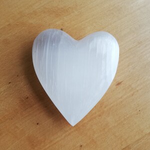 May include: A white heart-shaped selenite crystal with a smooth, polished surface. The crystal has a natural, striated texture.