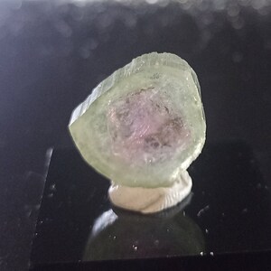 Könnte beinhalten: Ein durchscheinender grüner und rosa Kristall mit abgerundeter Form. Der Kristall liegt auf einer weißen Basis.