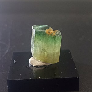 May include: A green and yellow crystal with a rough, natural texture. The crystal is displayed on a black surface.