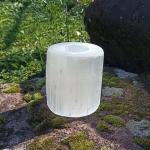 May include: White selenite candle holder with a smooth, polished surface. The holder is cylindrical in shape and has a hollow center for a candle. It is sitting on a mossy rock.