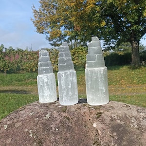 May include: Three white selenite crystal towers of varying sizes, arranged on a gray rock. The towers are made of stacked, layered crystals, giving them a unique and geometric appearance.