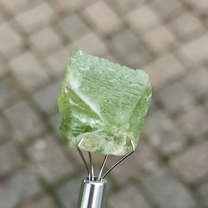 Puede incluir: Una piedra preciosa de peridoto verde claro, tallada en bruto, sujeta por una herramienta de metal plateado. El peridoto tiene una estructura cristalina con algunas inclusiones blancas y marrones. La piedra preciosa se muestra sobre un fondo borroso de tonos neutros.