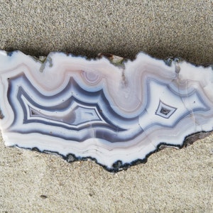 May include: A slice of agate with a white, gray, and pink banded pattern. The agate has a natural, rough edge.