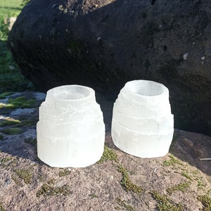 May include: Two white selenite candle holders, each with a unique, layered, and textured design. The candle holders are sitting on a mossy rock surface.
