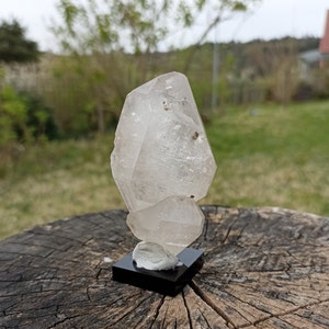 May include: A clear quartz crystal cluster with multiple points, displayed on a black stand. The crystal is translucent and has a natural, rough texture.