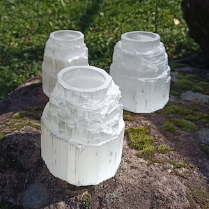 May include: Three white selenite candle holders, each with a unique, layered, crystal-like design. The holders are arranged on a rock surface with moss.