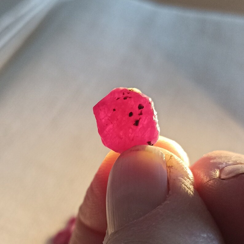 May include: A vivid, raw ruby gemstone, held between fingers. The ruby is a deep, saturated pink-red colour with dark inclusions. The gemstone has a rough, natural texture and is illuminated by soft light.