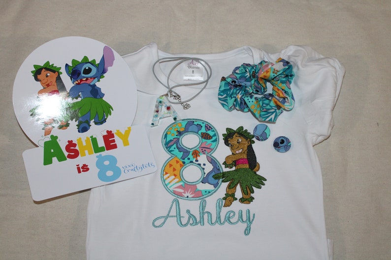 Puede incluir: Una camiseta blanca con un n&uacute;mero 8 bordado en azul y verde, un personaje de Stitch bordado en azul y verde, y el nombre "Ashley" bordado en azul. La camiseta forma parte de un conjunto de fiesta de cumplea&ntilde;os que incluye una coleta azul y verde, un collar azul y verde con un dije de la letra A, y un letrero redondo blanco con el texto "Ashley tiene 8 a&ntilde;os" y una caricatura de Stitch y Lilo.