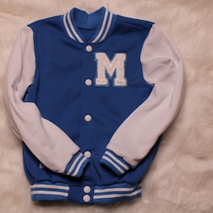 May include: A blue and white varsity jacket with a white "M" on the front. The jacket has white sleeves and blue and white stripes on the cuffs and bottom.