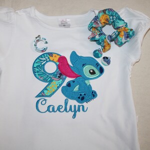 Puede incluir: Camiseta blanca con un personaje azul Stitch y el n&uacute;mero 9 bordado. El n&uacute;mero 9 est&aacute; rodeado de hojas y flores tropicales. El nombre Caelyn est&aacute; bordado debajo del personaje.