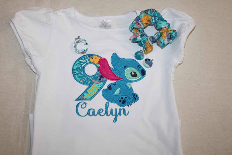 Puede incluir: Camiseta blanca con un personaje azul Stitch y el n&uacute;mero 9 bordado. El n&uacute;mero 9 est&aacute; rodeado de hojas y flores tropicales. El nombre Caelyn est&aacute; bordado debajo del personaje.