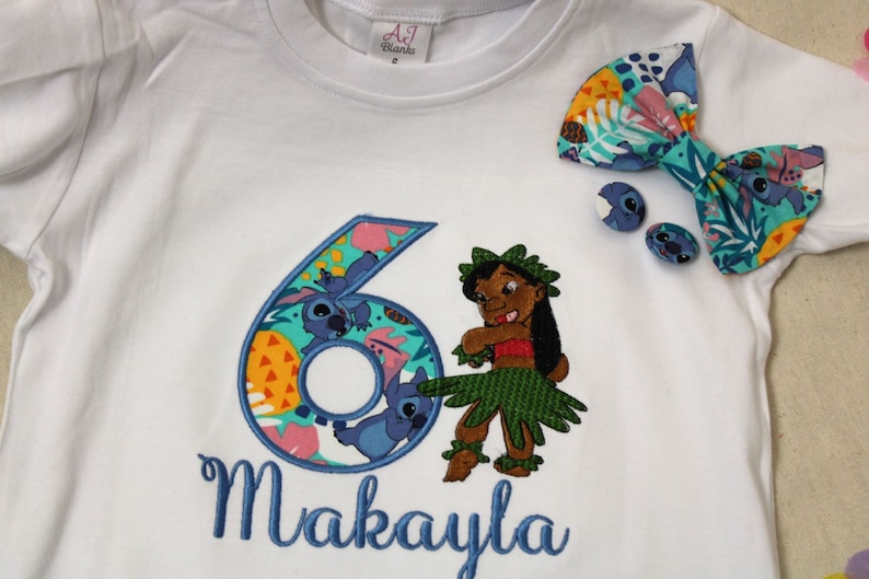 Puede incluir: Camiseta blanca con un n&uacute;mero seis bordado en azul, un personaje de dibujos animados y el nombre Makayla. El n&uacute;mero seis est&aacute; rodeado de un patr&oacute;n tropical colorido. Un lazo azul con un patr&oacute;n tropical est&aacute; en el hombro derecho de la camiseta.