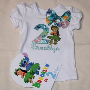 Puede incluir: Camiseta blanca con un n&uacute;mero 2 azul y verde, un lazo floral y el nombre "Brooklyn" bordado. Un personaje azul y verde a juego est&aacute; en la camiseta. Un cartel separado dice "Brooklyn tiene 2 a&ntilde;os".