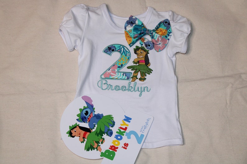 Puede incluir: Camiseta blanca con un n&uacute;mero 2 azul y verde, un lazo floral y el nombre "Brooklyn" bordado. Un personaje azul y verde a juego est&aacute; en la camiseta. Un cartel separado dice "Brooklyn tiene 2 a&ntilde;os".