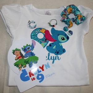 Puede incluir: Una camiseta blanca con un personaje azul Stitch bordado en la parte delantera. La camiseta dice "Caly is 9" y tiene un personaje azul y verde Stitch en un c&iacute;rculo blanco separado. Hay una goma para el pelo azul y verde y un amuleto azul y verde.