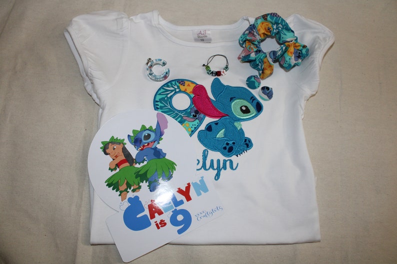 Puede incluir: Una camiseta blanca con un personaje azul Stitch bordado en la parte delantera. La camiseta dice "Caly is 9" y tiene un personaje azul y verde Stitch en un c&iacute;rculo blanco separado. Hay una goma para el pelo azul y verde y un amuleto azul y verde.