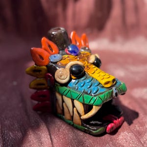 Puede incluir: Una colorida escultura artesanal de cabeza de dragón. El dragón presenta un diseño detallado con vibrantes tonos de azul, amarillo, naranja y verde. La escultura tiene dientes afilados y una boca abierta detallada.
