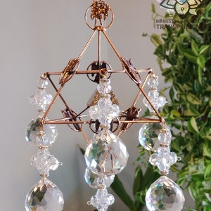 Pode incluir: Um apanhador de sol geométrico de arame de cobre com um design de estrela de David e múltiplos prismas de cristal pendurados nele. O apanhador de sol está pendurado no teto e está rodeado de plantas verdes.