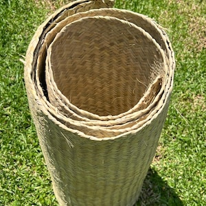 Pode incluir: Um tapete cilíndrico tecido à mão, feito de folhas de palmeira secas ou erva de cor castanho-claro. O tapete está enrolado firmemente em espiral, exibindo um padrão de tecido em espinha consistente em toda a sua superfície e bordas.