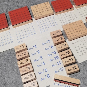 May include: A collection of wooden rubber stamps featuring calendar grids, month names, and dates. The stamps have light wood blocks and red rubber bases. Several stamped impressions on paper show monthly calendars and lists of months from Jan to Dec.