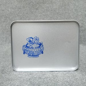 Op de afbeelding: Een rechthoekig zilverkleurig metalen blik met afgeronde hoeken. Het deksel is voorzien van een blauwe grafische print van een decoratieve urn gevuld met bloemen, met ingewikkelde geometrische patronen op de voet en twee kleine handvatten.
