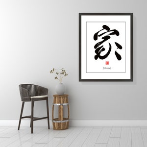 May include: A black framed print with a white background and a black calligraphy symbol in the center. The symbol is the Chinese character for "home". The word "Home" is written in black below the symbol in square brackets. The print is hanging on a white wall with a wooden stool and a chair in front of it.