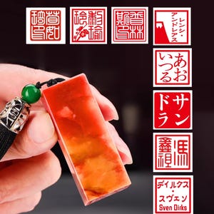Könnte beinhalten: Ein rechteckiges, orange-rotes Steinsiegel mit schwarzer Quaste und grüner Perle. Im Hintergrund befinden sich mehrere rote quadratische Stempel mit weißem Text. Der Text auf den Stempeln ist auf Japanisch und in anderen Sprachen.