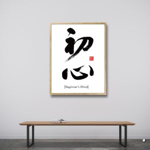 Pu&ograve; includere: Una stampa incorniciata con calligrafia nera su sfondo bianco. La calligrafia recita "Beginner's Mind" in inglese sotto i caratteri. La cornice &egrave; in legno chiaro.