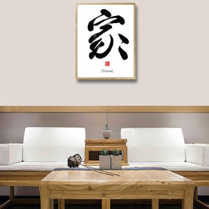 May include: A framed black and white calligraphy print of the Chinese character for "home" with a red seal and the word "Home" in English below it. The print is hanging on a light beige wall above a wooden coffee table and a white sofa.