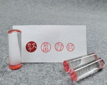 May include: Clear acrylic stamps with red ink pads. The stamps are cylindrical with rounded ends. A white card with four red Japanese characters is behind the stamps. The background is a gray fabric.