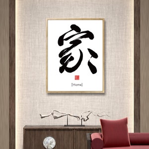 May include: A framed black and white calligraphy artwork with the Chinese character for "home" in a traditional style. The character is written in black ink on a white background. The frame is a simple wooden frame with a light brown finish. The artwork is hanging on a wall with a beige textured surface. The word "Home" is written in English below the character.