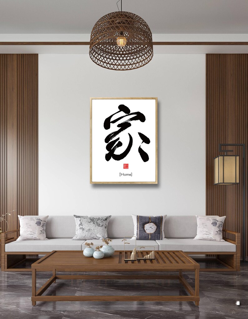 May include: A framed black and white calligraphy print of the Chinese character for "home" with the English translation in a red square below. The print is hanging on a white wall in a modern living room with a brown wooden sofa and coffee table.
