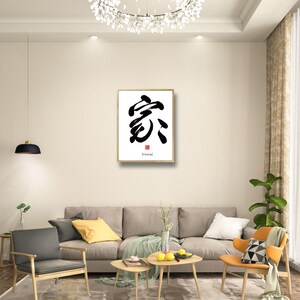 May include: A framed calligraphy artwork featuring the Chinese character for "home" in black ink on a white background. The character is written in a bold, brushstroke style. The frame is gold and the artwork is hanging on a white wall in a living room setting.