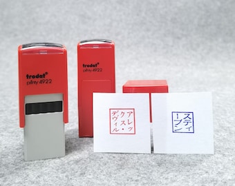 May include: Two red and gray Trodat Printy 4922 self-inking stamps. One stamp is upright, the other is lying down. Two white paper squares show stamped impressions: one in red ink with Japanese text in a square, and one in blue ink with Japanese text.