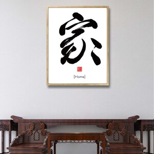 May include: A framed black and white calligraphy artwork of the Chinese character for "home" with the English translation "[Home]" below it. The artwork is hanging on a white wall above a wooden table and chairs.