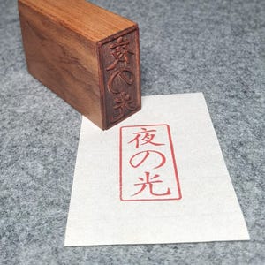 Puede incluir: Un sello de madera con caracteres japoneses tallados en un lado. El sello tiene forma rectangular y está hecho de madera oscura. Un papel muestra una impresión de tinta roja de los mismos caracteres japoneses.