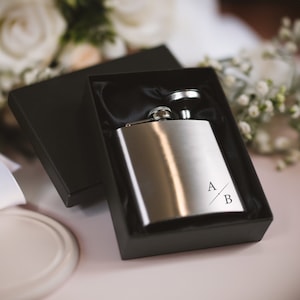 May include: A silver flask in a black box with a black satin lining. The flask has the initials "A" and "B" engraved on it.