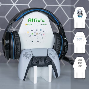 May include: White gaming console stand with a black controller and a black headset. The stand has a white background with a pixelated video game design and the text "Alfie's" and "43210".
