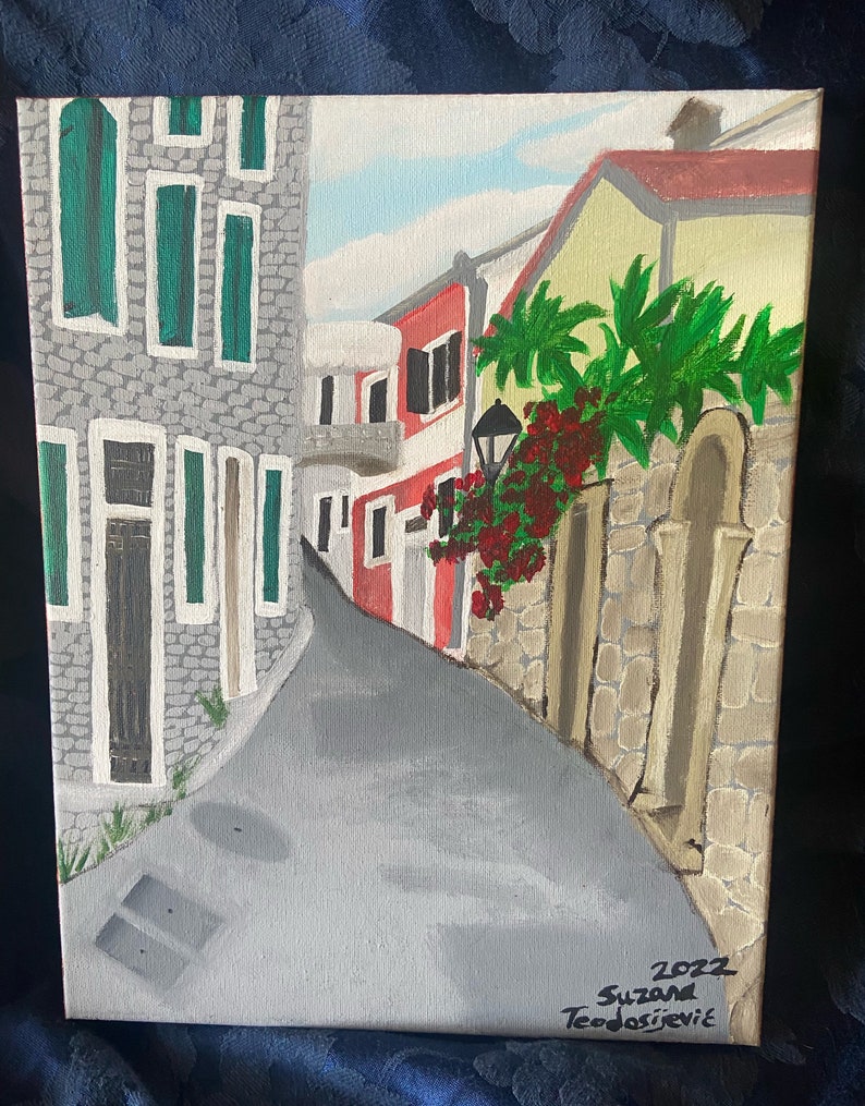 Puede incluir: Una pintura de una calle estrecha en un pueblo europeo. Los edificios est&aacute;n pintados en tonos de gris, rojo y amarillo. Hay plantas verdes y flores rojas creciendo a lo largo del lado de la calle. La pintura est&aacute; firmada "2022 Suzanna Teodossievic".
