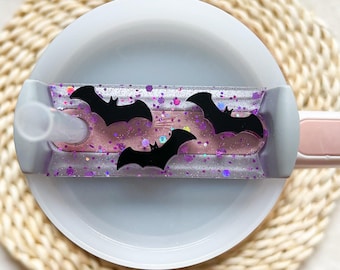 May include: A clear plastic cup with a purple and pink glitter design and black bat cutouts. The cup has a pink lid and a clear straw.