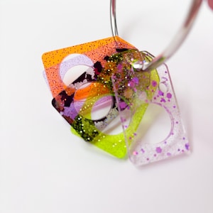 May include: A set of rectangular acrylic tags with circular cutouts, suspended on a silver metal ring. The tags feature translucent colors including orange, lime green, and clear, all embedded with various sizes of glitter in purple and gold tones.