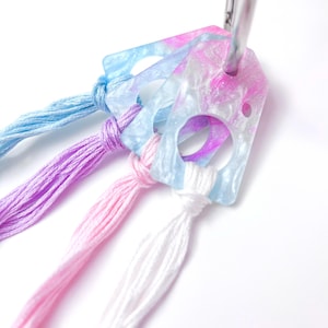 May include: A keychain with a clear, rectangular tag featuring a swirl of pink, purple, and blue colors. The tag has two circular cutouts and is attached to a silver ring. Colorful threads in blue, purple, pink, and white are knotted to the tag.