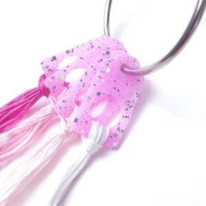 May include: A pink, glitter-speckled keychain with a silver ring. The keychain features a square design with cut-out shapes and is adorned with strands of pink, light pink, and white thread.