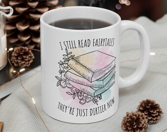 Può includere: Una tazza in ceramica bianca con un manico nero. La tazza ha un'illustrazione colorata di una pila di libri con il testo "I still read fairytales they're just dirtier now".