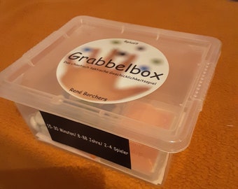 May include: A clear plastic storage box with a white label that reads "Grabbelbox" and "Das chaotisch taktische Geschicklichkeitsspiel" in German. The label also includes the name "René Borchers" and the text "15-30 Minuten/8-88 Jahre/2-4 Spieler".