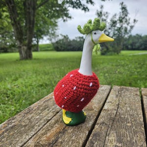 Puede incluir: Figura de ganso de cer&aacute;mica con cuello y cabeza blancos, pico amarillo y patas verdes y amarillas. El ganso lleva un disfraz de fresa de ganchillo rojo con detalles de semillas blancas y una corona de ganchillo verde. La figura est&aacute; sobre una superficie de madera.